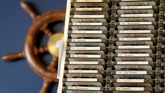 Kassetten mit den Titeln von Nationalhymnen stehen in einem Regal. Im Hintergrund ist ein Schiffssteuerrad zu sehen. © Zur redaktionellen Verwendung. Nicht-redaktionelle Verwendung nach Absprache. Foto: Marcus Brandt